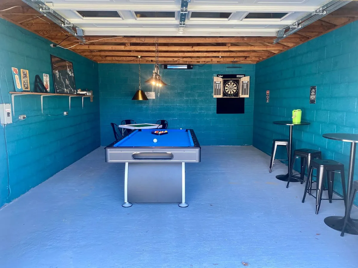This interior image features a game room setup with a pool table as the centerpiece, a dartboard on the wall, bar stools for casual seating, and a shelf with decorative items. The space invites guests to gather, play games, and socialize in a relaxed environment.