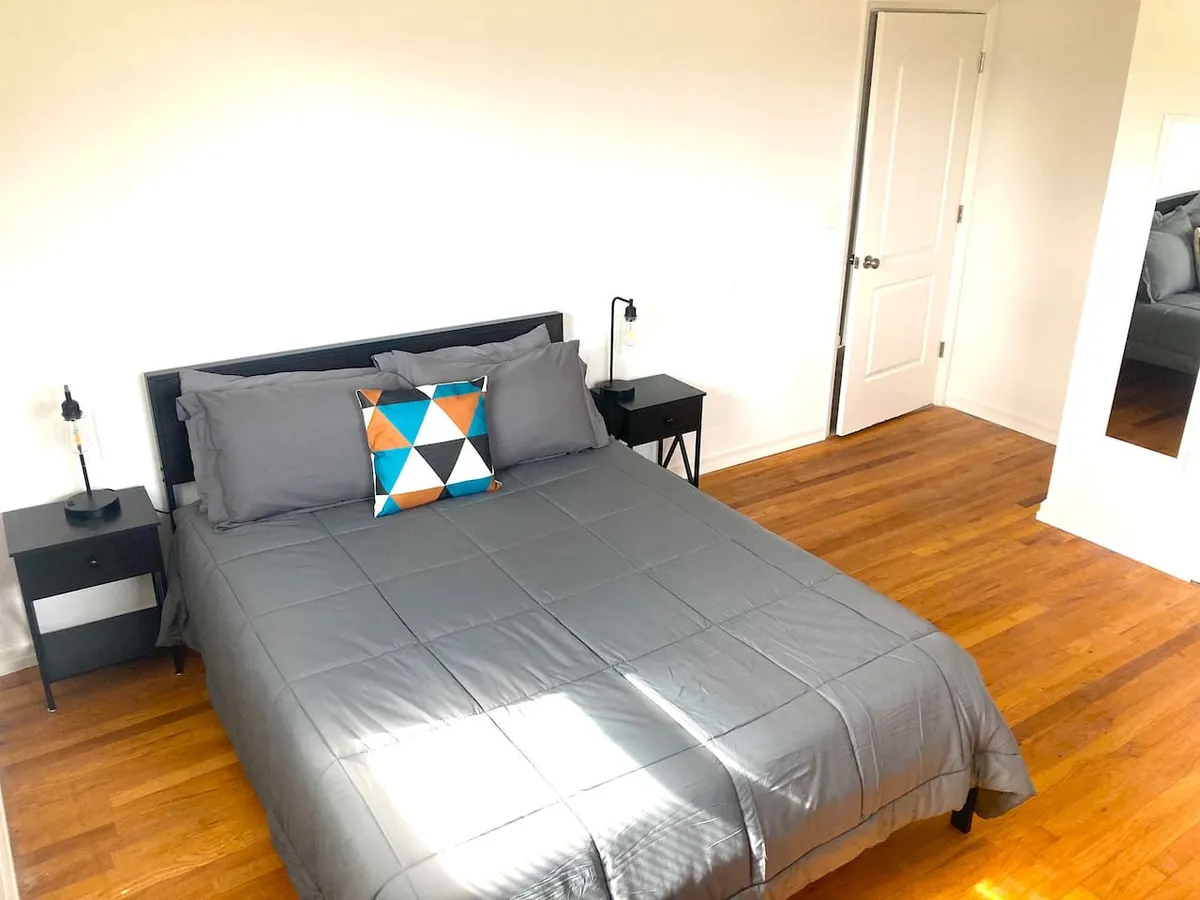 A tranquil bedroom highlighted by a bed with soft gray bedding, a colorful pillow, and nightstands with lamps casting a gentle glow. Natural light streams in, accentuating the serene and cozy atmosphere—perfect for unwinding or enjoying a restful night.