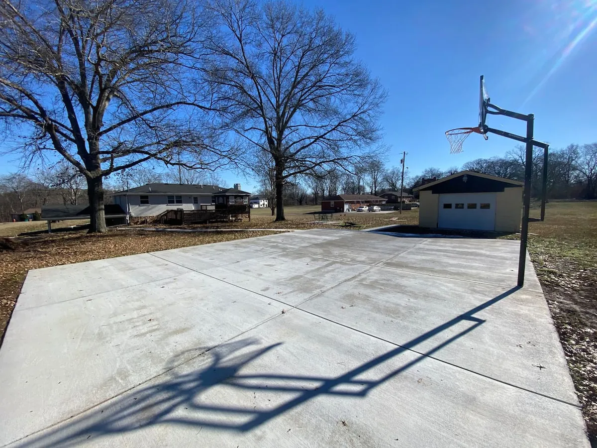 A vibrant outdoor scene featuring a private basketball court bordered by large trees, with a shed or garage visible in the background. The court is ready for pick-up games, and the surrounding lawn offers space for other outdoor activities, capturing an active, family-friendly atmosphere.