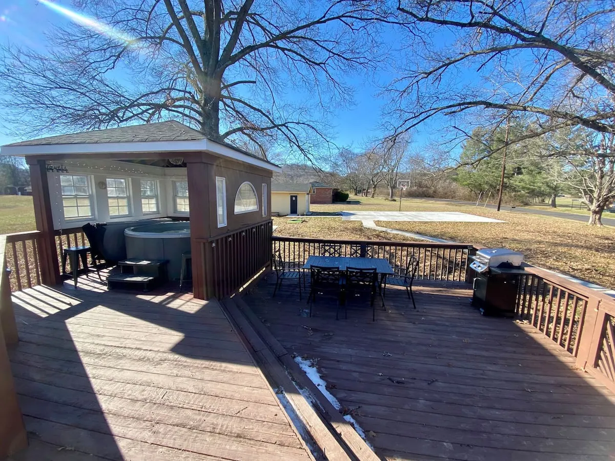 This vibrant exterior image showcases a spacious backyard featuring a bubbling hot tub, a large outdoor dining table set for meals, a BBQ grill ready for cookouts, and an open lawn area perfect for games. Sunlight pours over the scene, inviting guests to relax, dine, and play.