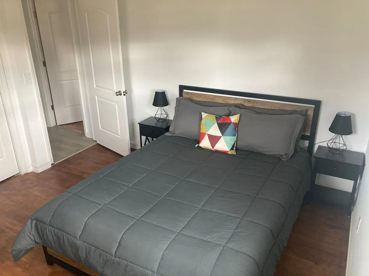 A serene bedroom featuring a neatly made bed with gray bedding, a colorful accent pillow, and nightstands with lamps on either side. The overall ambiance is welcoming, cozy, and restful—ideal for peaceful sleep after a long day. The space is uncluttered, with soft lighting that enhances the tranquil mood.
