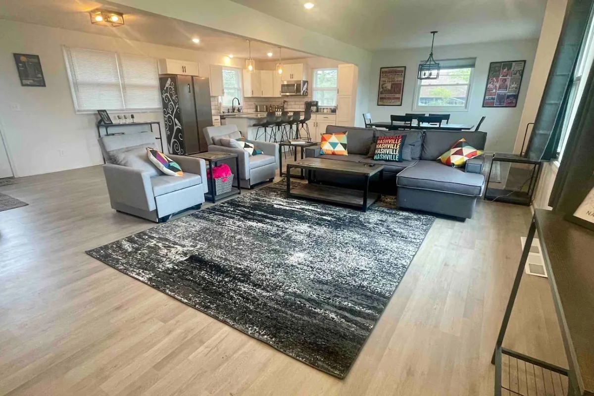 An open-plan living area showing a plush sofa with colorful cushions, a large area rug, a modern dining area with stylish chairs, and a kitchen island with bar stools. The layout invites both relaxation and communal dining, while the design balances comfort and style.