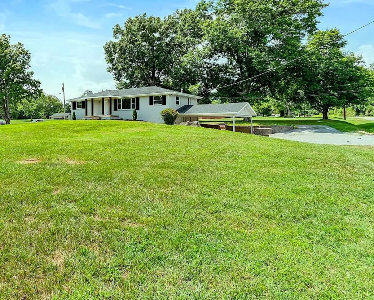 This image captures the exterior of the house, set against an expansive green lawn with mature surrounding trees. The open space offers ample room for pets and owners to relax or play, while the house is positioned invitingly in the background, hinting at the welcoming nature of the property.