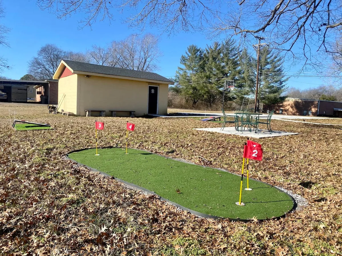 A playful and inviting exterior scene with a putting green complete with flags, an outdoor seating area perfect for gatherings, and a basketball hoop visible in the background. The yard is designed for friendly competitions, games, and relaxation.