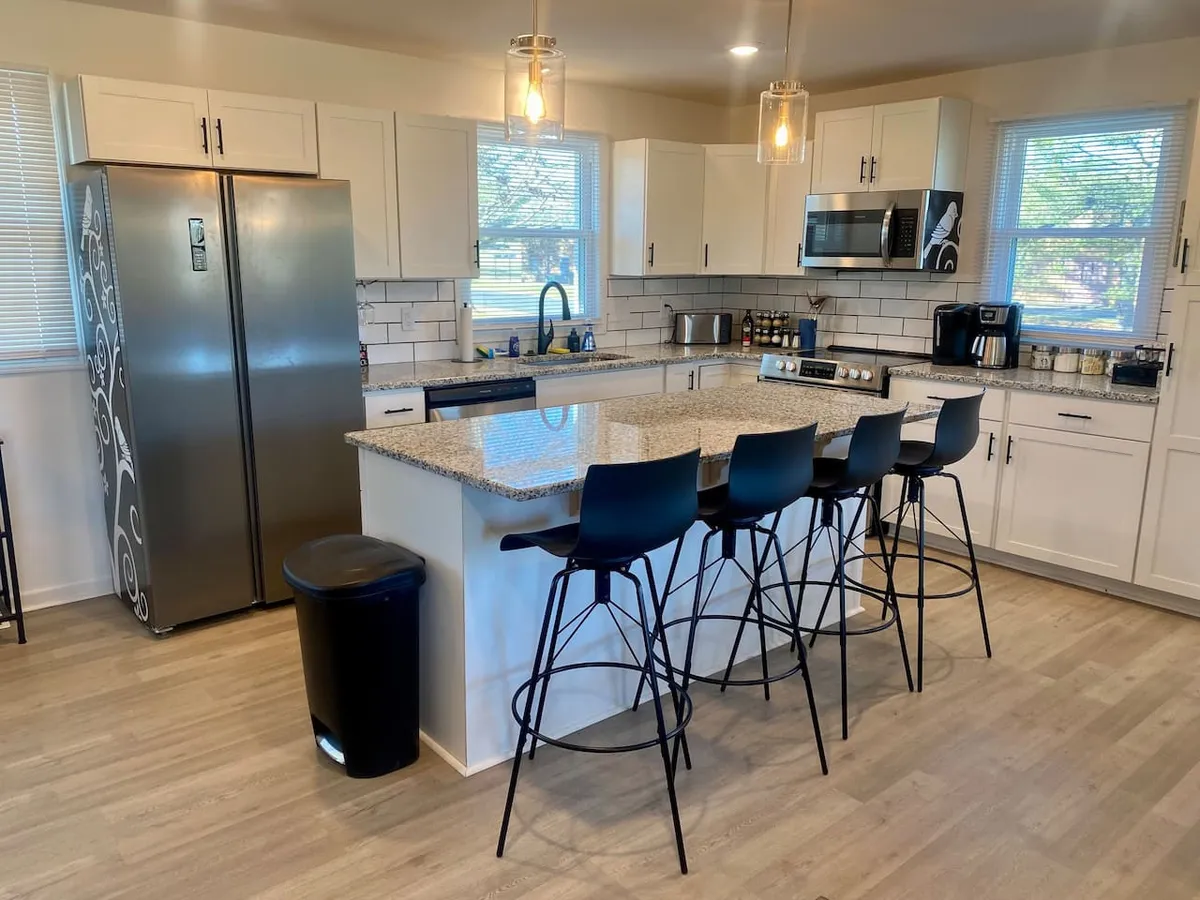A bright, open kitchen with sleek granite countertops, modern bar stools, stainless steel refrigerator, stove, and contemporary kitchen cabinets. The space is filled with natural light and invites group cooking and socializing, with ample room for movement and conversation.