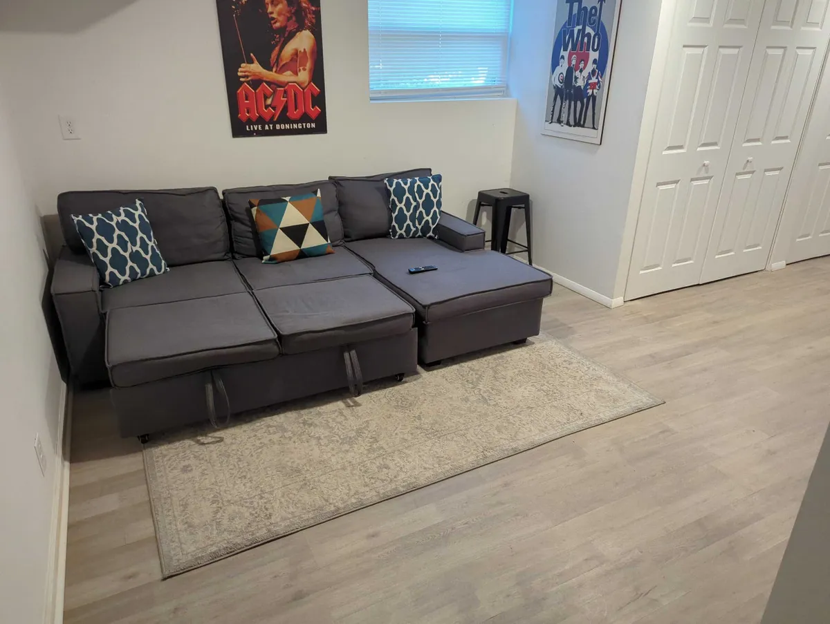 A comfortable living area featuring a sectional sofa, music-themed posters (AC/DC and The Who), a rug, and a small side table. The space is designed for relaxing, watching TV, or entertaining guests. Its casual ambiance and convertible furniture hint at its dual function as a lounge and a potential sleeping space for extra guests.
