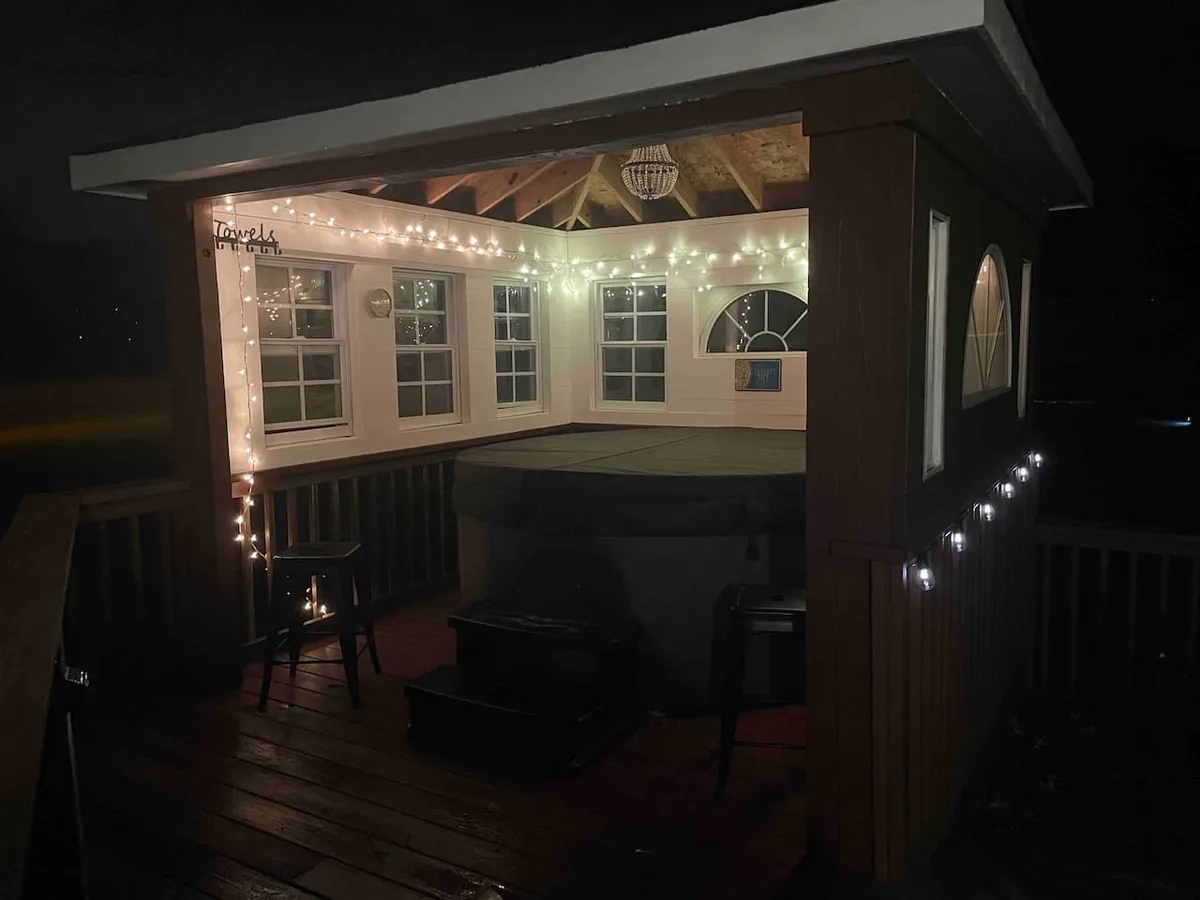 A cozy, intimate view of a wooden deck adorned with string lights and a decorative chandelier, centered around a steaming hot tub beneath the trees. The space glows with a magical ambiance, suggesting nighttime relaxation and group gatherings under the stars.