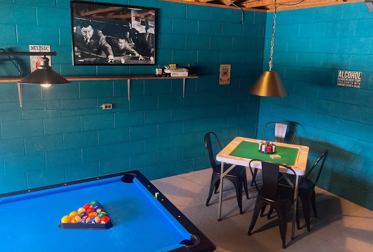 The image showcases a spacious, inviting interior with a stylish pool table as the focal point, a cozy dining area perfect for games or drinks, framed wall art adding local character, and a decorative shelf lending warmth. The scene suggests a lively yet relaxed atmosphere ideal for groups to gather, play, and unwind together.